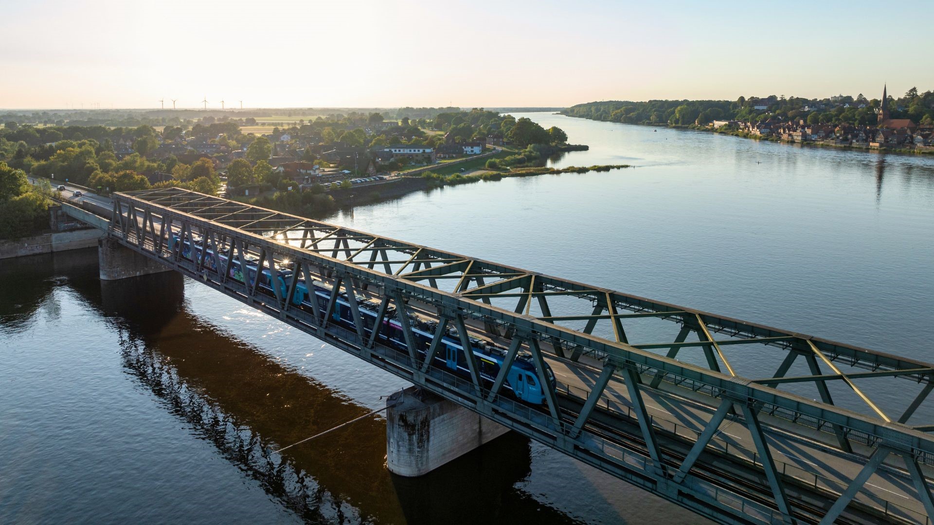 Eisenbahnbrücke über einen breiten Fluss bei Sonnenaufgang, mit einem blauen Zug auf der Brücke und grüner Landschaft im Hintergrund.