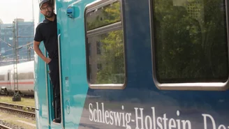 Person steht in der geöffneten Tür eines blau-türkisen Regionalzugs mit der Aufschrift ‚Schleswig-Holstein. Der echte Norden.‘ auf einem Gleisbereich, im Hintergrund Oberleitungen und ein weiterer Zug.