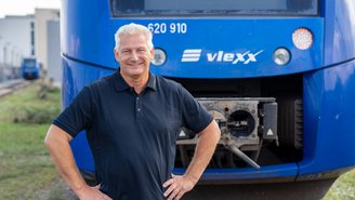 Person steht vor einem blauen vlexx-Zug auf Gleisen und trägt ein schwarzes Polo-Shirt