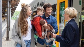 Familie mit Kind und Stofftier steht am Bahnsteig vor einem grünen und weißen Zug der vogtlandbahn und spricht mit einer Zugbegleiterin.