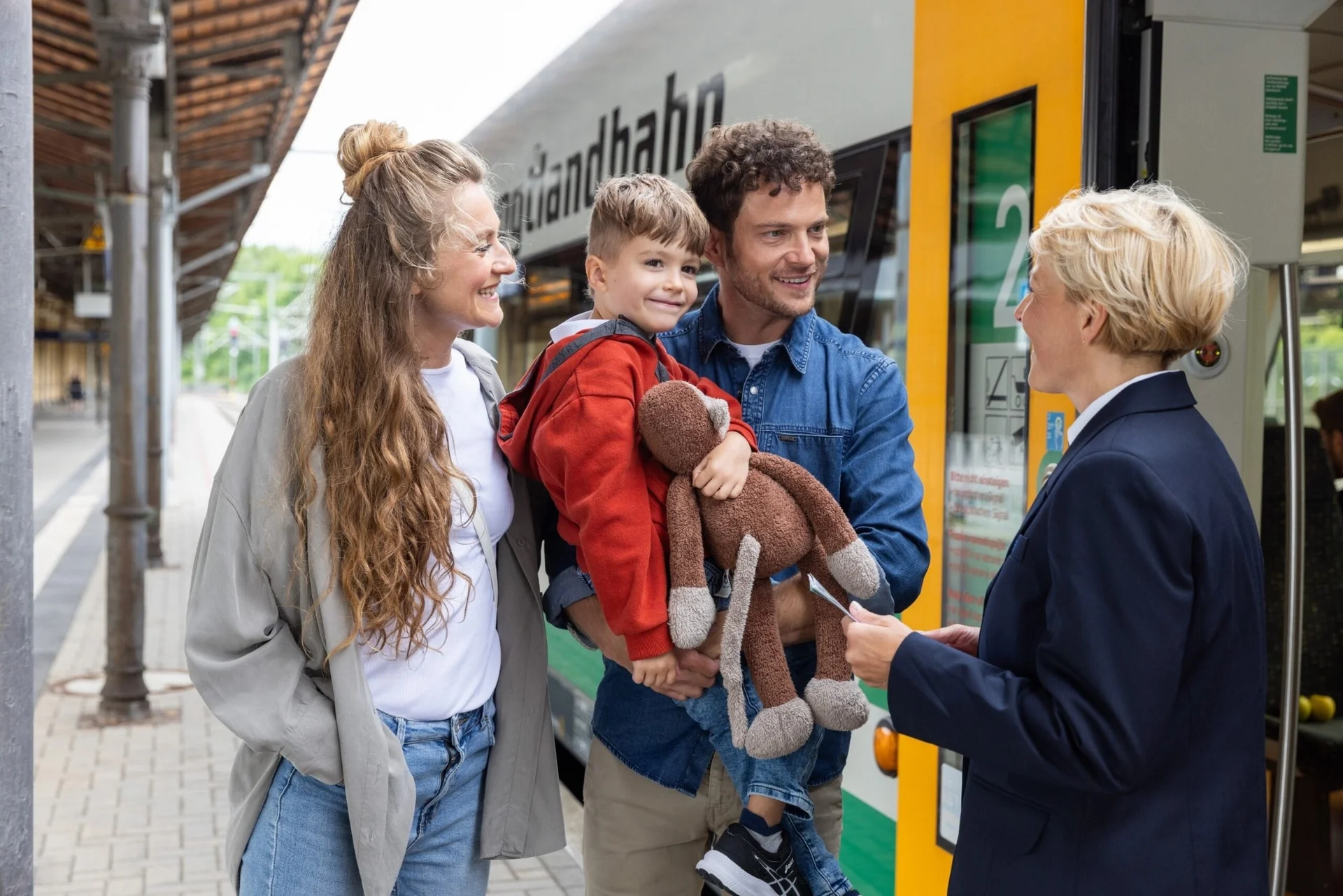 Familie mit Kind und Stofftier steht am Bahnsteig vor einem grünen und weißen Zug der vogtlandbahn und spricht mit einer Zugbegleiterin.