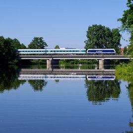Moderner blau-weißer Zug überquert eine Brücke über einen ruhigen Fluss, umgeben von grünen Bäumen und spiegelndem Wasser unter klarem Himmel.