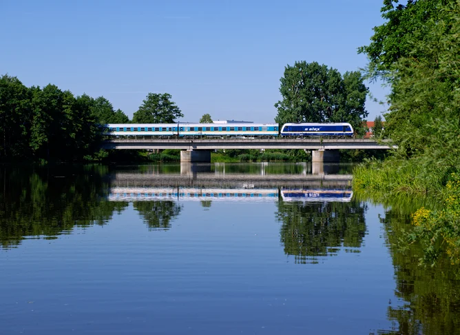 Moderner blau-weißer Zug überquert eine Brücke über einen ruhigen Fluss, umgeben von grünen Bäumen und spiegelndem Wasser unter klarem Himmel.
