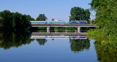 Moderner blau-weißer Zug überquert eine Brücke über einen ruhigen Fluss, umgeben von grünen Bäumen und spiegelndem Wasser unter klarem Himmel.