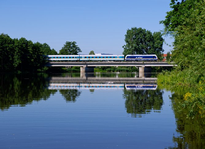 Moderner blau-weißer Zug überquert eine Brücke über einen ruhigen Fluss, umgeben von grünen Bäumen und spiegelndem Wasser unter klarem Himmel.