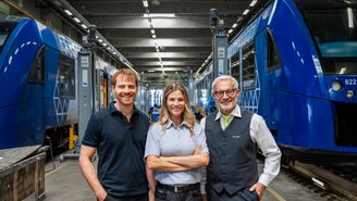 Drei Personen in Arbeitskleidung stehen in einer modernen Werkstatthalle zwischen zwei blauen vlexx-Zügen