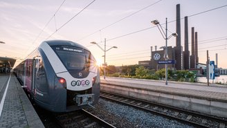 Moderner Regionalzug der Marke enno steht am Bahnsteig des Bahnhofs Wolfsburg, im Hintergrund das Volkswagen-Werk mit Schornsteinen bei Sonnenuntergang.