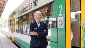 Person in dunkelblauer Dienstuniform steht mit verschränkten Armen vor einem grünen und weißen Zug der vogtlandbahn am Bahnsteig.