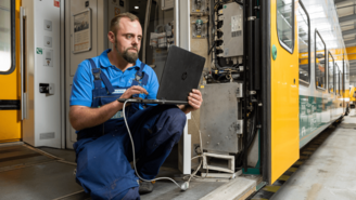 Person in blauer Arbeitskleidung arbeitet mit einem Laptop an elektronischen Komponenten im geöffneten Schaltschrank eines gelb-grünen Zuges in einer Werkstatt