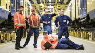 Fünf Personen in Arbeitskleidung stehen in einer Werkstatthalle zwischen zwei gelb-blauen Zügen, einer liegt entspannt mit Klemmbrett auf dem Boden