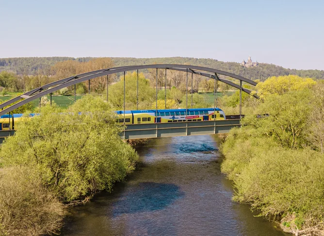 Gelb-blauer Regionalzug überquert eine Stahlbrücke über einen Fluss, umgeben von grüner Landschaft und Feldern unter klarem blauem Himmel.