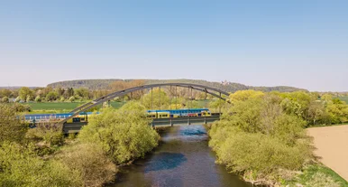 Gelb-blauer Regionalzug überquert eine Stahlbrücke über einen Fluss, umgeben von grüner Landschaft und Feldern unter klarem blauem Himmel.