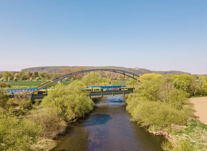 Gelb-blauer Regionalzug überquert eine Stahlbrücke über einen Fluss, umgeben von grüner Landschaft und Feldern unter klarem blauem Himmel.