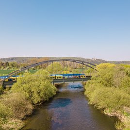 Gelb-blauer Regionalzug überquert eine Stahlbrücke über einen Fluss, umgeben von grüner Landschaft und Feldern unter klarem blauem Himmel.