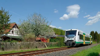 Grün-weißer Regionalzug der vogtlandbahn fährt auf einer eingleisigen Strecke durch eine ländliche Landschaft mit Fachwerkhaus, blühendem Baum und blauem Himmel.