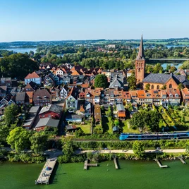 Blau-türkiser Regionalzug fährt entlang eines Flusses vor einer historischen Altstadt mit roten Dächern, Kirche und Schloss, umgeben von grüner Landschaft und blauem Himmel.