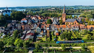 Blau-türkiser Regionalzug fährt entlang eines Flusses vor einer historischen Altstadt mit roten Dächern, Kirche und Schloss, umgeben von grüner Landschaft und blauem Himmel.