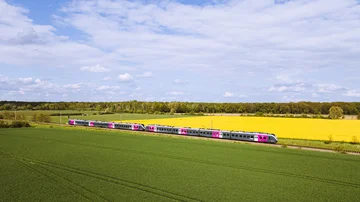 Silberner Regionalzug mit pinken Akzenten fährt durch eine grüne Landschaft mit Feldern und gelbem Raps unter blauem Himmel mit Wolken, aufgenommen aus der Luft.
