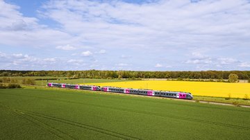 Silberner Regionalzug mit pinken Akzenten fährt durch eine grüne Landschaft mit Feldern und gelbem Raps unter blauem Himmel mit Wolken, aufgenommen aus der Luft.