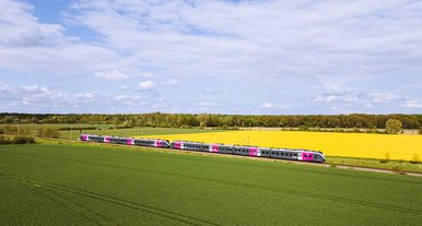 Silberner Regionalzug mit pinken Akzenten fährt durch eine grüne Landschaft mit Feldern und gelbem Raps unter blauem Himmel mit Wolken, aufgenommen aus der Luft.