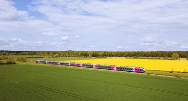 Silberner Regionalzug mit pinken Akzenten fährt durch eine grüne Landschaft mit Feldern und gelbem Raps unter blauem Himmel mit Wolken, aufgenommen aus der Luft.
