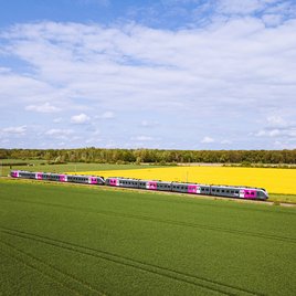 Silberner Regionalzug mit pinken Akzenten fährt durch eine grüne Landschaft mit Feldern und gelbem Raps unter blauem Himmel mit Wolken, aufgenommen aus der Luft.