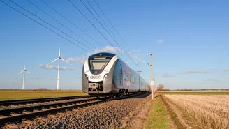 Moderner Elektrozug fährt durch offene Landschaft mit Windkraftanlagen im Hintergrund, klare blaue Himmel und Schienen im Vordergrund.