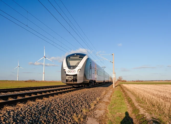 Moderner Elektrozug fährt durch offene Landschaft mit Windkraftanlagen im Hintergrund, klare blaue Himmel und Schienen im Vordergrund.