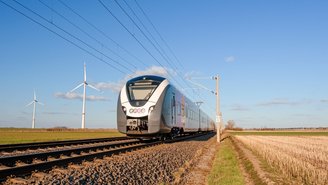 Moderner Elektrozug fährt durch offene Landschaft mit Windkraftanlagen im Hintergrund, klare blaue Himmel und Schienen im Vordergrund.