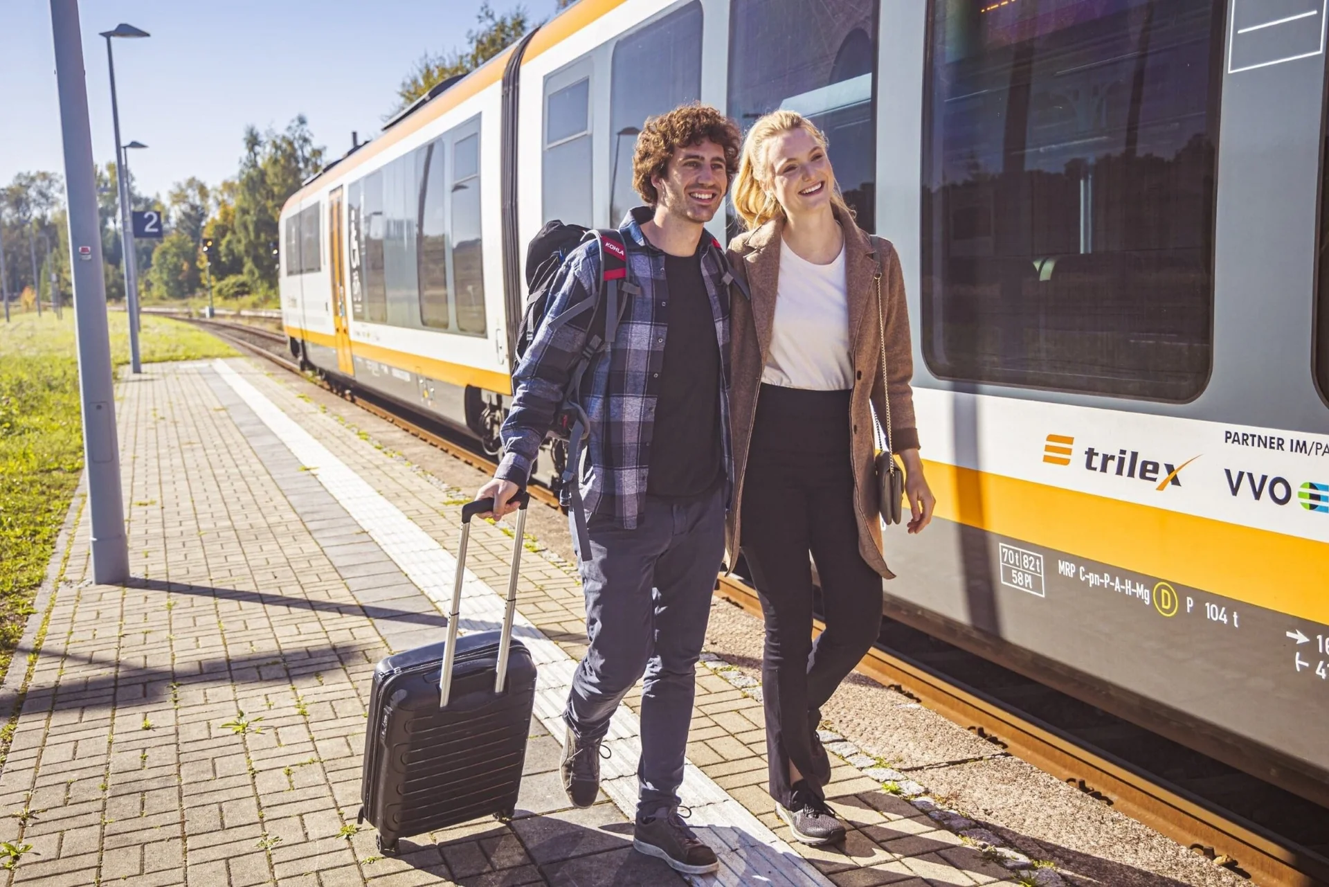 Zwei Reisende stehen auf einem Bahnsteig neben einem orange-weißen Regionalzug der Marke trilex, einer zieht einen Rollkoffer.