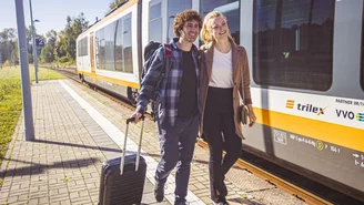 Zwei Reisende stehen auf einem Bahnsteig neben einem orange-weißen Regionalzug der Marke trilex, einer zieht einen Rollkoffer.