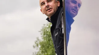 Zugpersonal lehnt sich aus dem geöffneten Fenster eines modernen Zuges, vor einem Hintergrund mit Bäumen und bewölktem Himmel.