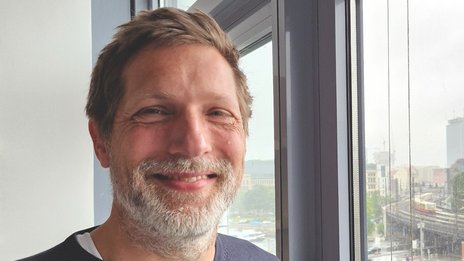 Christian Canis, Leiter Bahnbetriebsplanung bei NETINERA Deutschland GmbH, in einem Büro mit großem Fenster und Blick auf Bahninfrastruktur und Stadtlandschaft.