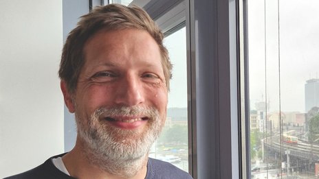 Christian Canis, Leiter Bahnbetriebsplanung bei NETINERA Deutschland GmbH, in einem Büro mit großem Fenster und Blick auf Bahninfrastruktur und Stadtlandschaft.