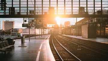 „Leerer Bahnhof Zoologischer Garten mit Gleisen und moderner Glasfassade bei Sonnenaufgang, Lichtstrahlen spiegeln sich auf dem Bahnsteig.