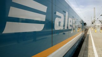 Blauer Regionalzug mit der Aufschrift ‚alex‘ steht am Bahnsteig eines modernen Bahnhofs, Spiegelung des Logos auf der Zugseite sichtbar.