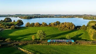 Blau-türkiser Regionalzug fährt entlang eines Sees durch eine grüne Landschaft mit Wiesen und Bäumen unter klarem Himmel, aufgenommen aus der Luft.