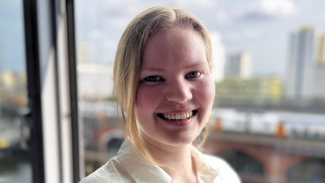 Lucie Girod, Werkstudentin in der Betriebsplanung bei NETINERA Deutschland GmbH, in einem Büro mit großem Fenster und Blick auf Fluss und Stadtarchitektur.