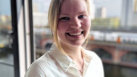 Lucie Girod, Werkstudentin in der Betriebsplanung bei NETINERA Deutschland GmbH, in einem Büro mit großem Fenster und Blick auf Fluss und Stadtarchitektur.