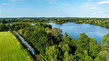 Blau-türkiser Regionalzug fährt entlang eines Sees durch eine grüne Landschaft mit Wäldern und Wiesen unter blauem Himmel, aufgenommen aus der Luft.
