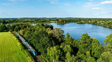 Blau-türkiser Regionalzug fährt entlang eines Sees durch eine grüne Landschaft mit Wäldern und Wiesen unter blauem Himmel, aufgenommen aus der Luft.