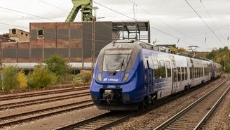 Blauer vlexx-Regionalzug fährt auf Gleisen vor einem Industriegebäude mit Förderturm im Hintergrund
