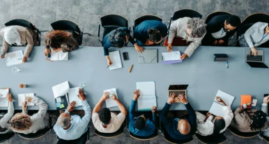 Große Gruppe von Personen sitzt an einem langen Konferenztisch und arbeitet gemeinsam an Dokumenten, Notebooks und Laptops in einem modernen Besprechungsraum