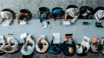 Große Gruppe von Personen sitzt an einem langen Konferenztisch und arbeitet gemeinsam an Dokumenten, Notebooks und Laptops in einem modernen Besprechungsraum