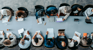 Große Gruppe von Personen sitzt an einem langen Konferenztisch und arbeitet gemeinsam an Dokumenten, Notebooks und Laptops in einem modernen Besprechungsraum