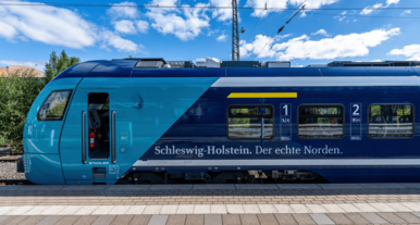 Blau-türkiser Regionalzug mit der Aufschrift ‚Schleswig-Holstein. Der echte Norden.‘ steht am Bahnsteig unter blauem Himmel mit vereinzelten Wolken.