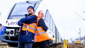 Zwei Personen in orangefarbenen Warnwesten stehen vor einem modernen erixx Regionalzug auf einem Gleisbereich, umgeben von Oberleitungen und Bahninfrastruktur.