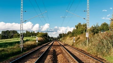 Doppelgleisige Eisenbahnstrecke mit Oberleitungen, umgeben von grüner Landschaft und klarem Himmel.