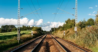 Doppelgleisige Eisenbahnstrecke mit Oberleitungen, umgeben von grüner Landschaft und klarem Himmel.