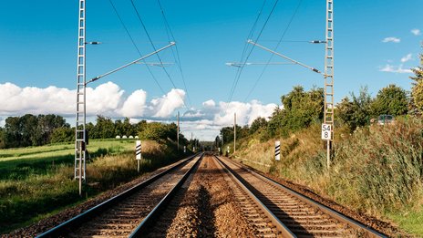 Doppelgleisige Eisenbahnstrecke mit Oberleitungen, umgeben von grüner Landschaft und klarem Himmel.